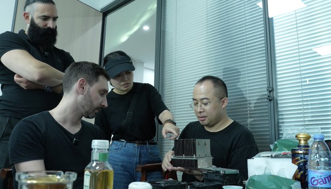 A group of four people in black clothing gathered around a table indoors, with one person holding what appears to be a piece of equipment.