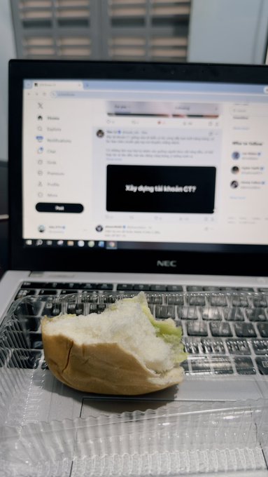 A casual photo of a partially eaten sandwich resting on the keyboard of an NEC laptop, with the screen displaying a social media feed with Vietnamese text including 'Xây dựng thị khoan CTTP'.