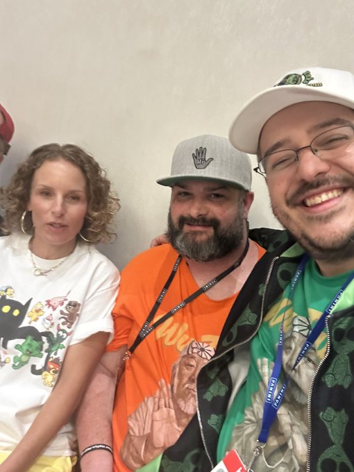 A selfie of three smiling people posing together, including a woman in a graphic tee, a bearded man in an orange shirt and gray cap, and a man in a green hoodie and glasses with lanyards.