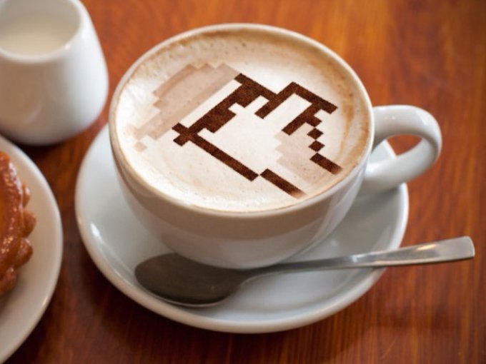 A cappuccino in a white cup and saucer featuring pixel art latte art depicting a Minecraft-style axe.