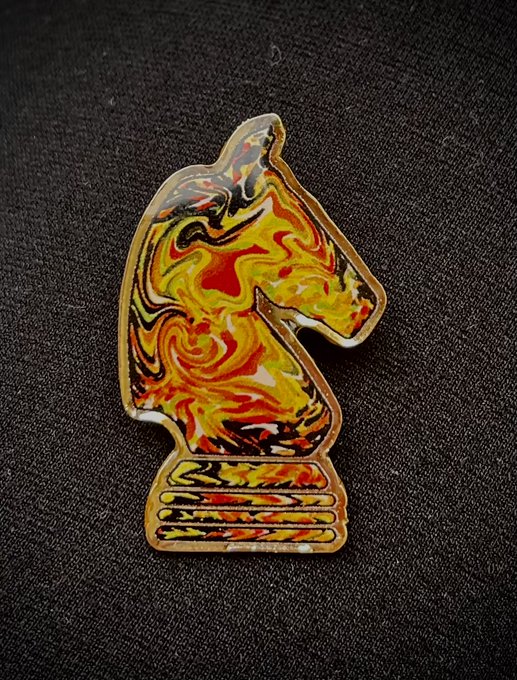 A vibrant, marbled enamel pin shaped like a chess knight, featuring swirling patterns of yellow, red, black, and white, rests on a dark fabric background—symbolizing the creator's first artistic mint from 2020 and a meaningful, limited keepsake they may share during their Singapore visit.