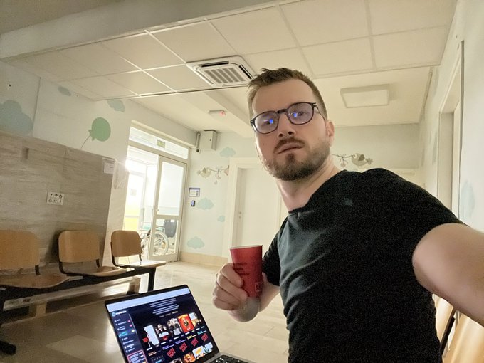A bearded man wearing glasses and a black t-shirt sits indoors holding a red coffee mug, with a laptop displaying a photo gallery on a table in front of him.