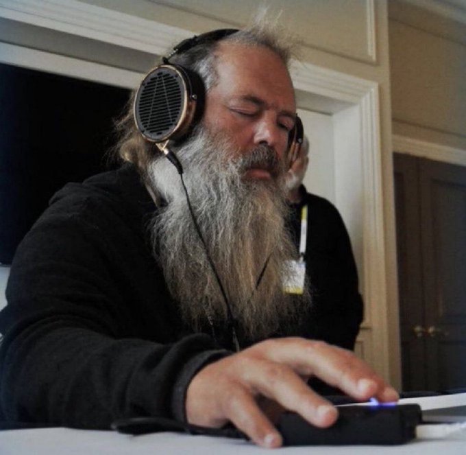 The image features a man with a long beard wearing headphones, sitting at a table with his hands on a gaming controller. The man appears to be playing video games or using some type of electronic device that requires physical interaction. There are two other people in the background who seem to be watching him play. The post about the Phantom bags suggests that the man might be involved in an online gaming community, where players often use virtual reality headsets and accessories like headphones for immersive gameplay experiences.