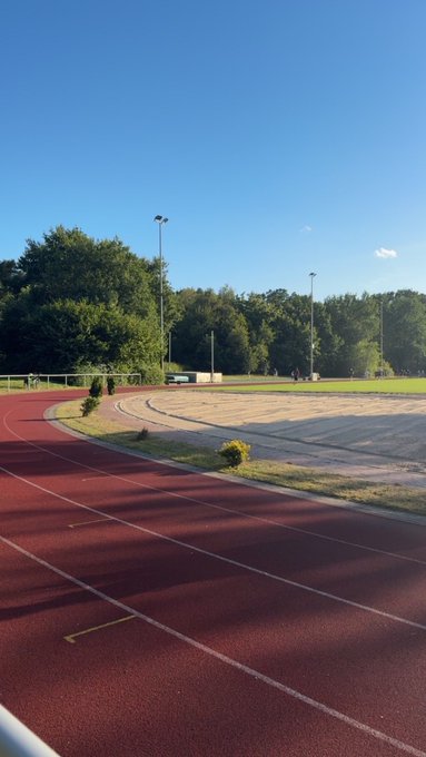 A running track with reddish-brown lanes curves around a green field under a clear blue sky, surrounded by trees and stadium lighting poles.