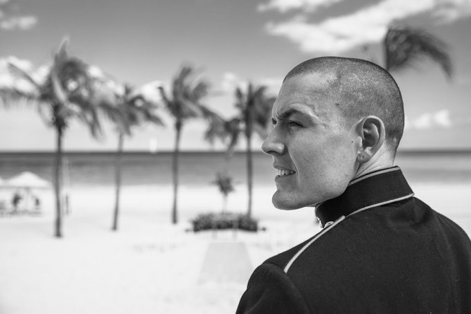A black-and-white photo shows a smiling, short-haired man in a dark jacket looking sideways, with palm trees and an ocean in the background.