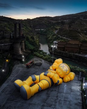 A large yellow inflatable character lies on a rooftop overlooking an industrial area with brick buildings and a river at dusk.