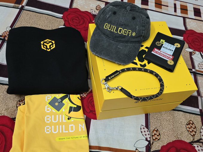 The image features a collection of merchandise items, including a black sweatshirt, a yellow shirt, a hat, and a book. The items are neatly arranged on a bedspread or blanket with red roses visible behind them. The text "builder" is prominently displayed in the center of the image, indicating that these items might be related to construction or building-related activities.