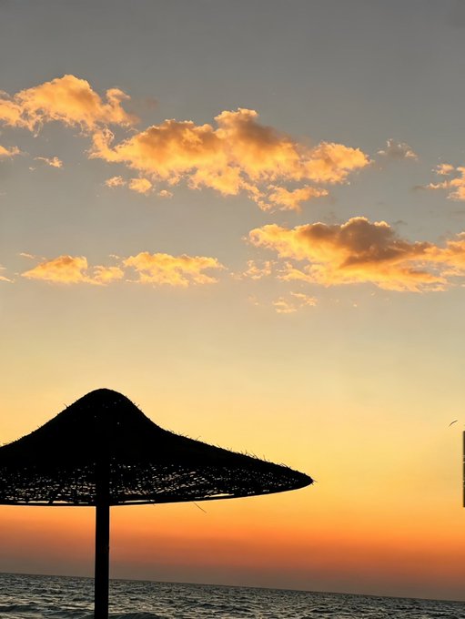 A scenic sunset photograph featuring a silhouetted straw beach umbrella against a golden sky with scattered clouds over the ocean.