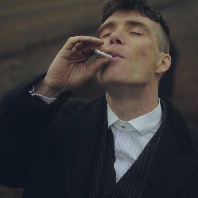 A cinematic shot of a man in a black coat and white shirt smoking a cigarette with his eyes closed.