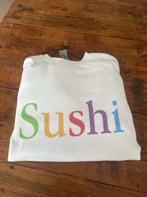 A folded white shirt on a wooden surface with the word 'Sushi' printed in colorful, rainbow letters (green, orange, pink, purple, blue).