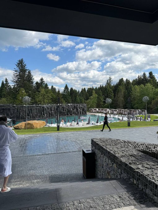 A scenic outdoor plaza with a waterfall feature, blue sky, and pine trees in the background, viewed from under a covered area where a person in a white robe stands.