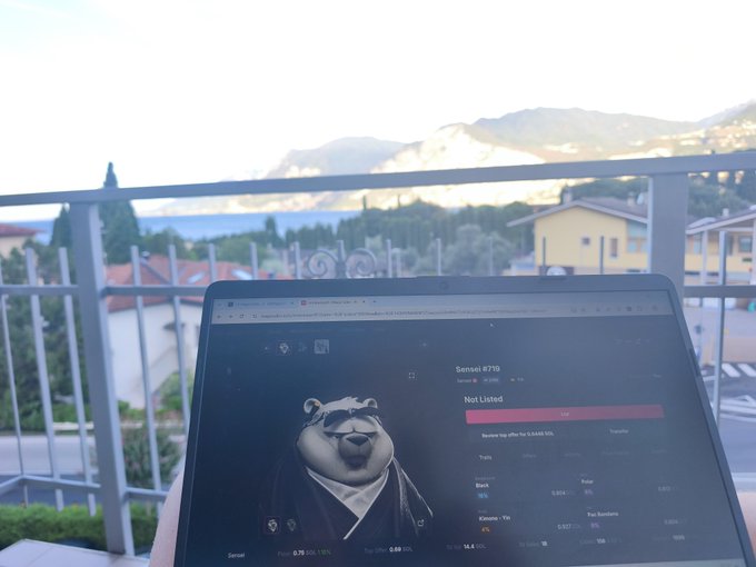 A laptop displaying a digital bear collectible (Sensei #719) being viewed on a balcony overlooking a mountain and lake landscape.