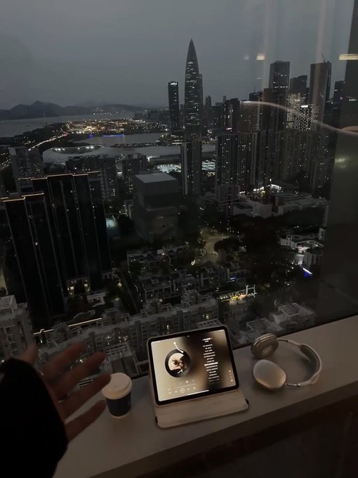 The image shows a person's hand reaching out towards an open window with a view of a city skyline at night. The scene includes a laptop, headphones, and a cup of coffee placed near the window. The background features tall buildings that illuminate the cityscape below. The text in the image reads "GM Some projects market products, @quipnetwork markets possibility. Stay qupinin...".