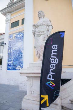 An outdoor scene featuring a 'Pragma' branded banner standing in front of a white statue of a seated woman, with blue and white decorative tiles (azulejos) visible on a building in the background.