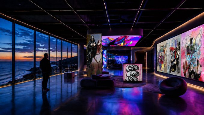 A solitary figure stands in a stunning, futuristic luxury home with floor-to-ceiling windows overlooking a coastal sunset, while vibrant digital artworks by Nodefnd illuminate the space's walls and ceiling, making it the ultimate immersive viewing experience.