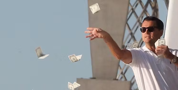 Leonardo DiCaprio in a white shirt and sunglasses throws money into the air against a blue sky.