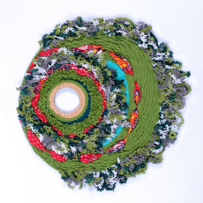 A circular textile artwork features a textured green knitted spiral adorned with small fabric trees and colorful patterned accents, resembling a miniature forest landscape.
