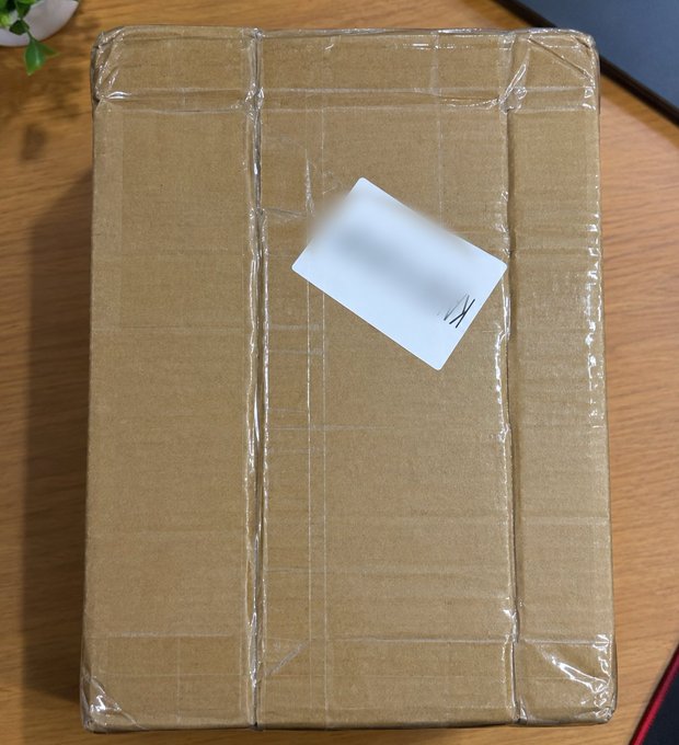 A sealed cardboard package wrapped in clear packing tape sits on a wooden table, featuring a white shipping label with the text blurred out except for a small handwritten letter.