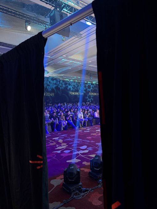 A view through stage curtains at TOKEN2049 shows a panel discussion underway with a packed audience seated in a large conference hall featuring ornate purple patterned carpeting and dramatic stage lighting. The glimpse from backstage perfectly captures the anticipation of the upcoming appearance by Mert, Balajis, and Cameron at the crypto conference.