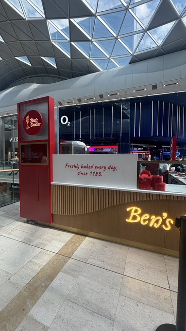 A Ben's Cookies kiosk in a shopping concourse near the O2 arena displays a sign reading "Freshly baked every day, since 1983." The post "GM Bros" suggests the founders of Ben's Cookies, who began selling their signature cookies in London in 1983.