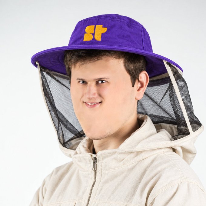 A product photo of a young man wearing a purple beekeeper-style hat with mesh veil and 'ST' logo in yellow, paired with a light beige hooded jacket.