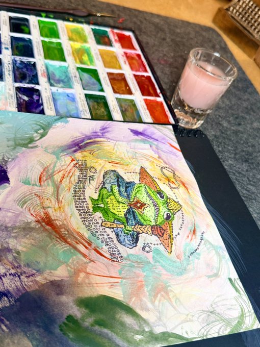 A colorful mixed-media artwork featuring a geometric green and orange creature sits beside a watercolor palette and a glass of pink liquid.