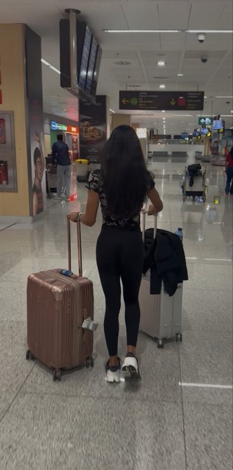 A woman with long dark hair, seen from behind, walking through an airport terminal while pulling a rose-gold suitcase and carrying a black bag.