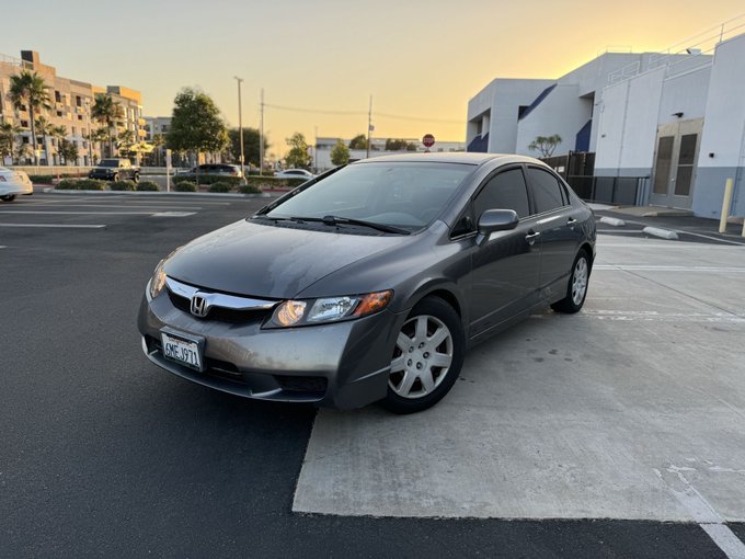 A gray Honda Civic sedan parked in a parking lot at sunset, with commercial buildings and a stop sign visible in the background.