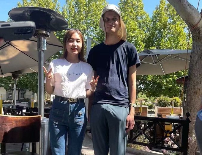 Two people pose outdoors at what appears to be an outdoor restaurant or cafe with umbrellas and patio furniture visible. The woman on the left wears a white t-shirt with 'Hether' text and high-waisted jeans with a Gucci belt, while the tall man on the right wears a black t-shirt, gray pants, and a sun hat with neck flap.