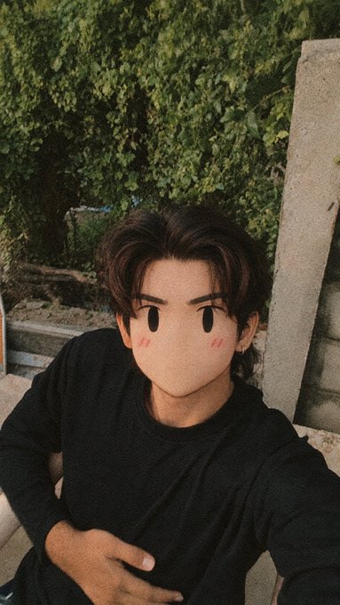 A selfie of a person outdoors with greenery in the background, whose face is obscured by a large cartoon-style Mii avatar sticker with rosy cheeks.
