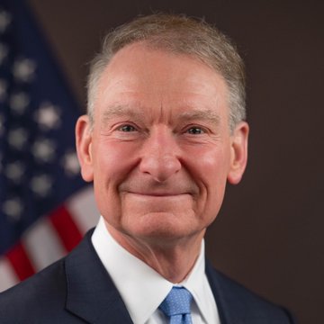 An official portrait of a gray-haired man in a dark suit and light blue tie, smiling in front of an American flag.