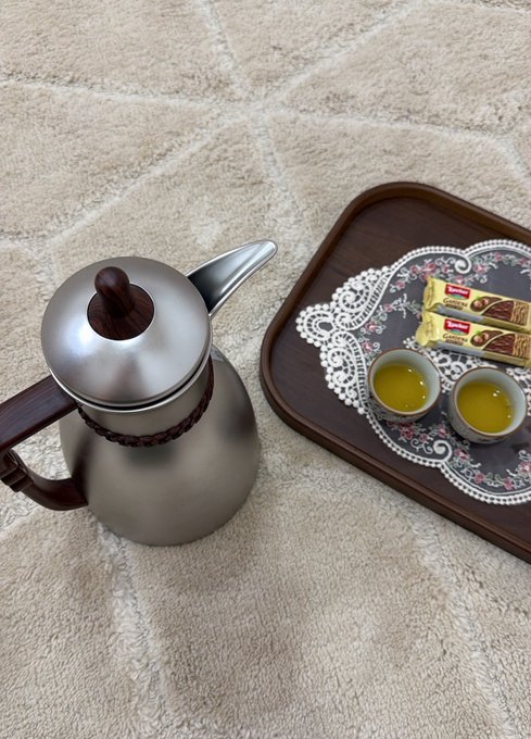 A stainless steel coffee pot with a wooden handle sits beside a decorative wooden tray holding two small cups of coffee and Lipton Yellow Label tea packets, all arranged on a beige patterned rug. The simple, intimate coffee setup reflects the post's celebration of understated luxury and a peaceful evening ritual.