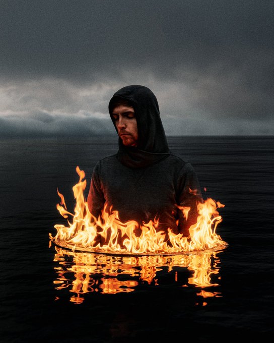 A surreal photograph of a man in a black hoodie sitting cross-legged in a dark ocean with his eyes closed in meditation, while a ring of fire burns on the water's surface around him.