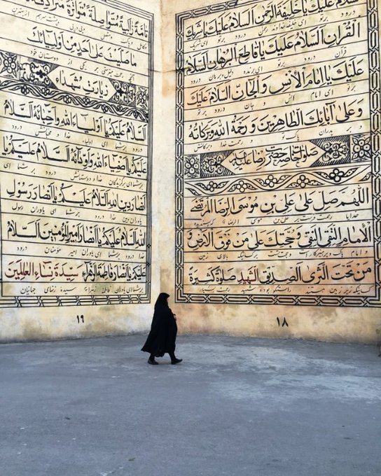 A solitary figure in a black chador walks across a concrete plaza in front of a massive, aged wall covered in large Arabic calligraphy panels marked with the numbers 18 and 19.