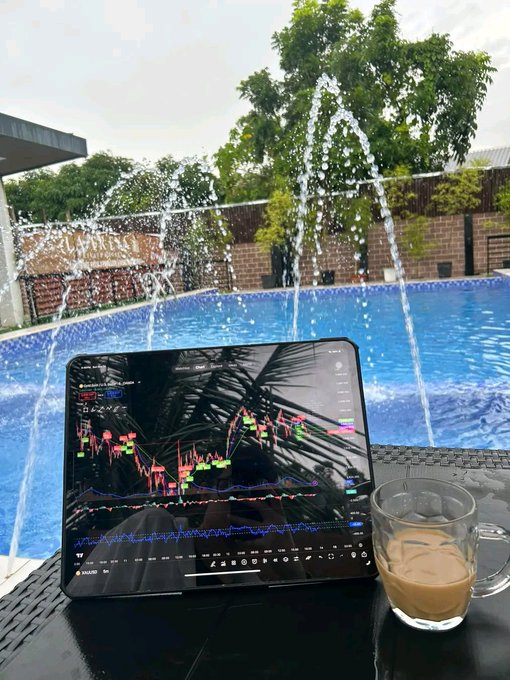 A tablet displaying a cryptocurrency trading chart with red and green candles sits on a black table next to a glass of iced coffee, with a blue swimming pool and water fountains in the background.