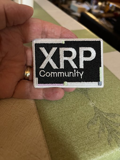 A hand holds an embroidered patch reading 'XRP Community' in white text on a black background, set against a wooden surface.