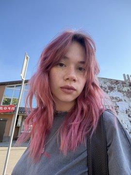 A woman with messy, pink hair takes a selfie against a blue sky, wearing a gray t-shirt and a black choker.