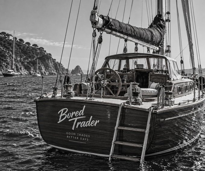 A black-and-white photograph of a sailboat named "Bored Trader" with the tagline "GOOD TRADES, BETTER DAYS" on its stern, featuring a boarding ladder and anchored near a coastline.