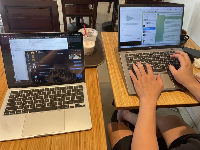 Two people sit side by side at a wooden table in a coffee shop, each typing on their respective laptops—one appears to be a MacBook running what looks like a Telegram or chat interface, while the other is a Dell laptop displaying a coding or development environment. A cold iced coffee with a red straw sits between them, perfectly capturing the "coffee session" mentioned in the post where they're chatting about coding, the "ARC" hot topic, and joining "Tempo."