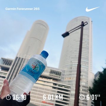 A hand holding a clear water bottle with a blue cap in front of tall modern skyscrapers, overlaid with Garmin Forerunner 265 and Nike branding and stats: 35:10, 6.01 km, 6'01".
