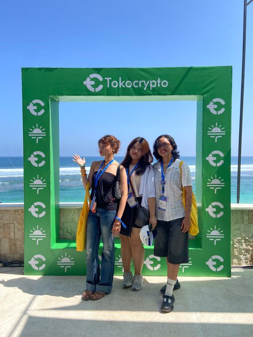 Three attendees pose at a beachside photo frame branded with the "Tokocrypto" logo, capturing a sunny seaside moment at the event.