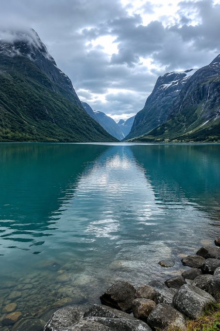 A scenic photograph of a calm, turquoise lake reflecting a cloudy sky, flanked by steep, green mountains with patches of snow, and rocks visible in the clear foreground water.