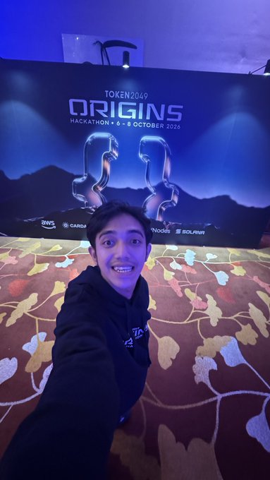 A selfie of a young man in a black hoodie at the TOKEN2049 ORIGINS Hackathon event (6-8 October 2024), with event branding and sponsor logos like AWS, CARDA, SOLVM visible in the background.