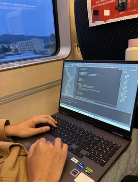 A person working on a laptop displaying code while riding on a train, with a window showing buildings passing by.