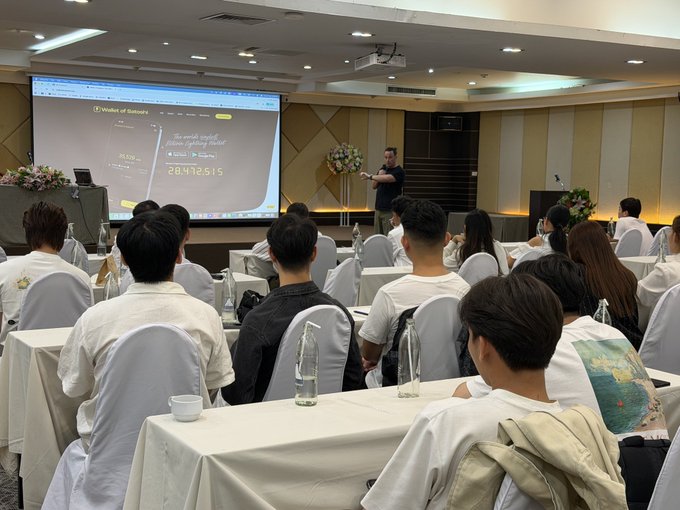 A presenter stands before an audience in a conference room displaying a slide titled "Wallet of Satoshi" with the number "29472515," delivering a talk that aligns with the post's note about on-boarding users to the @WalletofSatoshi platform.