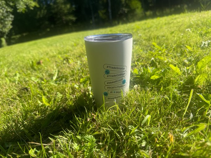 A white tumbler cup sitting in green grass with hashtags printed on it including #TradeSmarter, #TradeOnSnober, #MakeItCount, #DGBToTheMoon, and others related to cryptocurrency/trading.