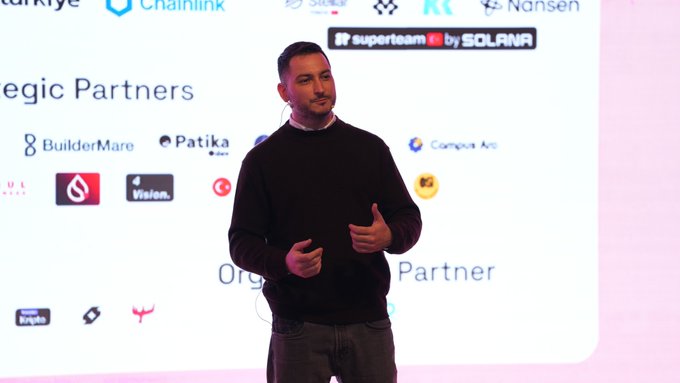 A man in a black sweater giving a presentation on stage with a backdrop listing sponsors including Chainlink, Solana Superteam, BuilderMare, Patika, and others.