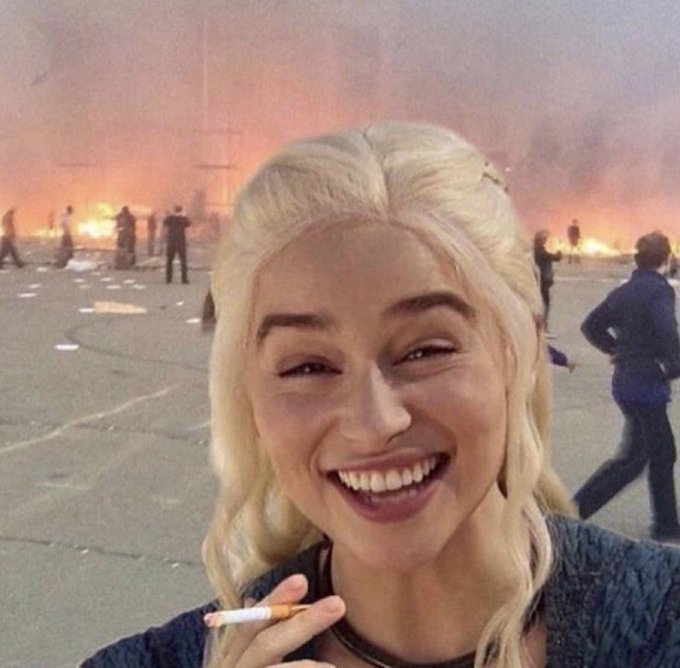 A blonde woman smiles widely while holding a cigarette in the foreground, with a chaotic scene of fires and crowds visible in the background.