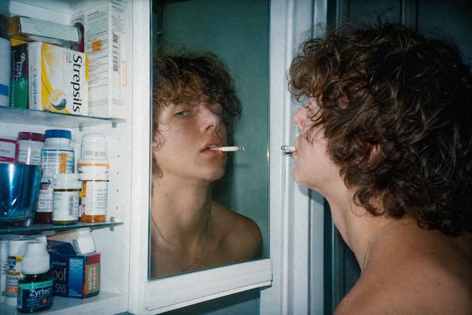 A young man with curly hair smokes a cigarette while looking at his reflection in a medicine cabinet mirror, with various prescription bottles and boxes like Strepsils visible on the shelves.