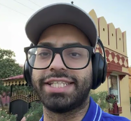 A selfie of a smiling man wearing a gray baseball cap, black glasses, and over-ear headphones, with a colorful structure visible in the background.